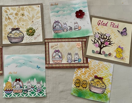 Sex färgglada påskkort med motiv av kaniner, kycklingar, ägg och blommor på en ljus duk.