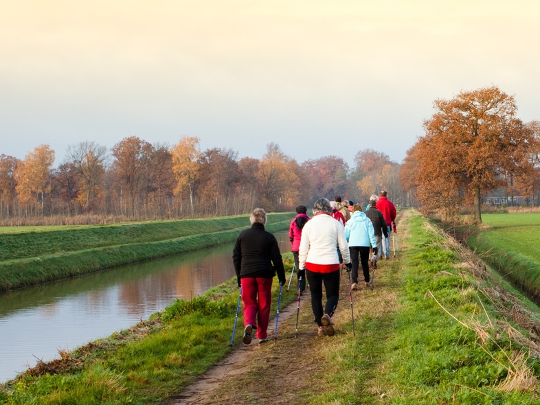 En grupp människor promenerar längs en kanal i ett höstlandskap med gröna fält och träd med orange löv.