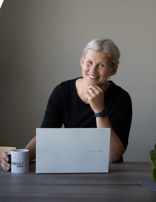 Leende kvinna med ljust hår sitter vid ett bord med en laptop och en kaffemugg.