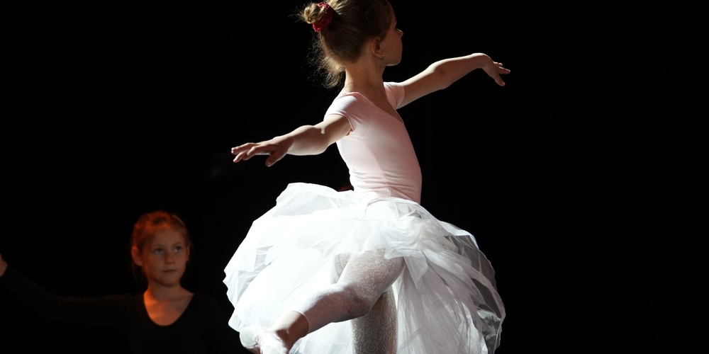 En ung ballerina i vit tyllkjol dansar på scenen med armarna utsträckta, belyst av scenljus.