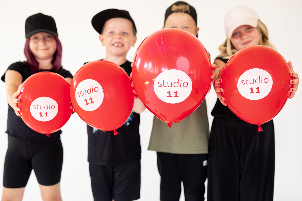 Fyra barn i svarta kläder håller röda ballonger med texten 'studio 11' mot kameran.