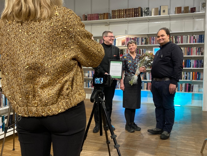 En kvinna fotograferar Per Lodenius, Lisa Förare och Quinten Moody som poserar i ett bibliotek, en av dem håller en blombukett.