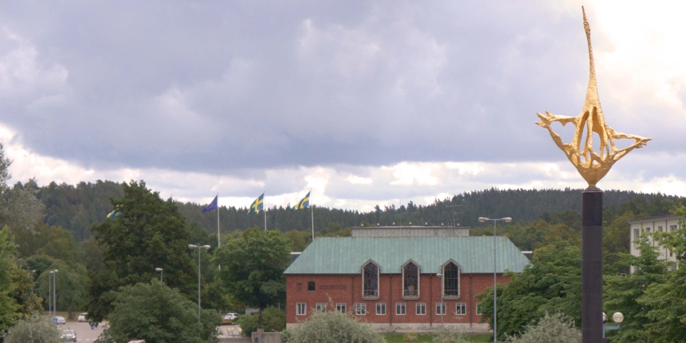 Stadsvy med en guldfärgad skulptur i förgrunden, grönska och en tegelbyggnad i bakgrunden.