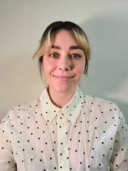 Kvinna med blont hår och prickig vit skjorta ler mot kameran framför en ljus bakgrund.