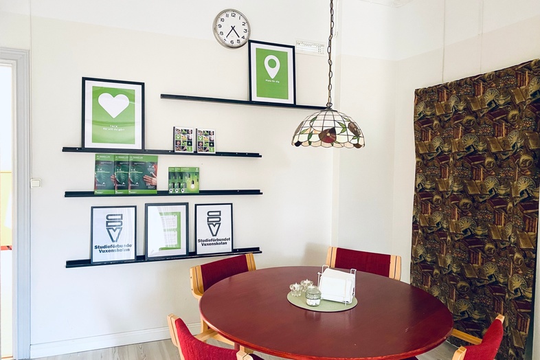 Ett ljust rum med ett runt bord, fyra röda stolar, väggklocka, hyllor med tavlor och en taklampa.