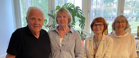 Fyra äldre personer står tillsammans inomhus framför stora fönster och en grön växt.