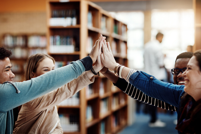 Fyra unga personer gör en gemensam high five i ett bibliotek, glada och engagerade.