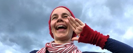 Person med röd mössa, randig halsduk och röda handledsvärmare poserar utomhus mot en molnig himmel.