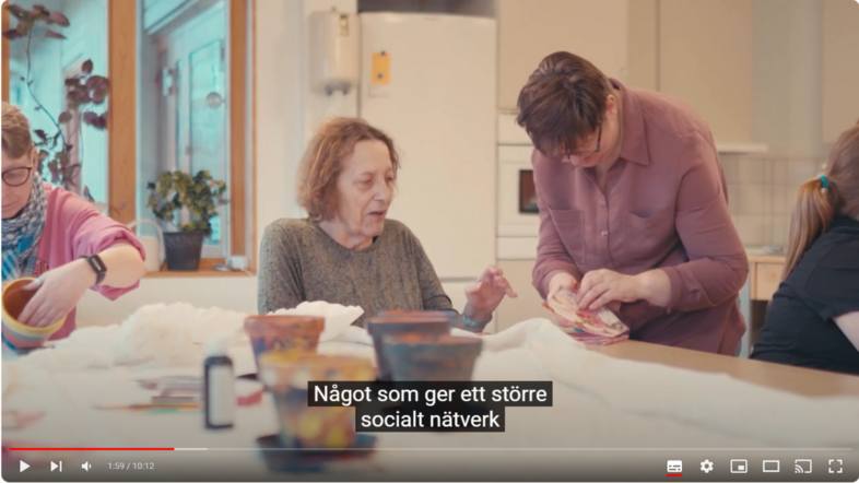 Tre kvinnor sitter vid ett bord och pysslar tillsammans i ett ljust rum. Svensk text visas på skärmen.