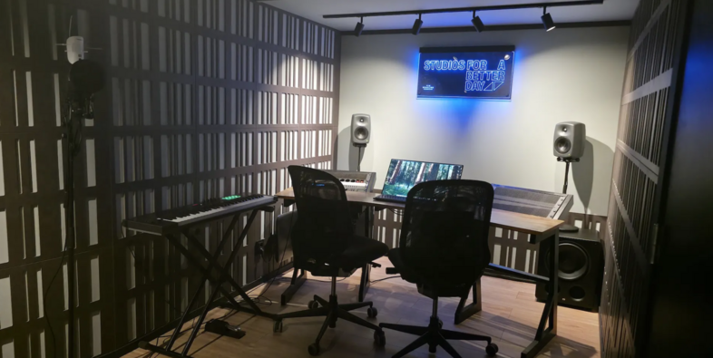 Ett modernt musikproduktionsrum med skrivbord, två stolar, högtalare, keyboard och en neonskylt på väggen.