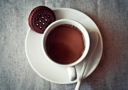 En kopp varm choklad på ett vitt fat med en chokladkaka och en vit sked, ovanifrån.