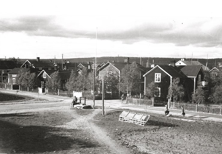 Svartvit bild av en liten by med trähus, grusvägar och några människor som promenerar.