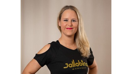 Kvinna med långt blont hår och svart t-shirt med texten 'Jallabina Workout' poserar mot beige bakgrund.