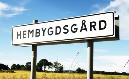 Vägskylt med texten 'Hembygdsgård' mot en lantlig bakgrund med blå himmel och fält.
