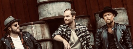 Tre personer sitter avslappnat framför staplade oljefat mot en trävägg, klädda i läderjackor och hattar.