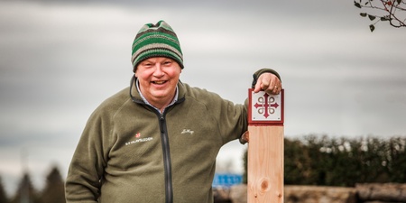 Person med grön mössa och grön jacka står utomhus och lutar sig mot en trästolpe med pilgrimsledssymbol.