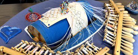 Närbild av en spetsknypplingsuppsättning med trådar, bobbiner och mönster på en blå kudde.