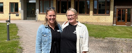 Två kvinnor står utomhus framför en byggnad med skylten 'BIBLIOTEK' under en blå himmel.