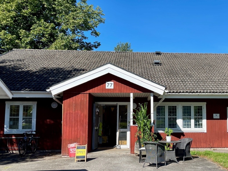 En låg faluröd byggnad med vita fönsterfoder och knutar och siffran 77 över entrén. I förgrunden en grusad uppfart, trädgårdsmöbler och krukväxt. I bakgrunden blå himmel och höga gröna lövträd.