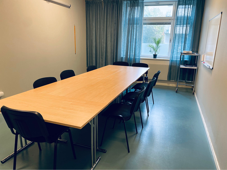 Ett tomt mötesrum med ett långt bord, åtta stolar, fönster med gardiner och en whiteboard på väggen.