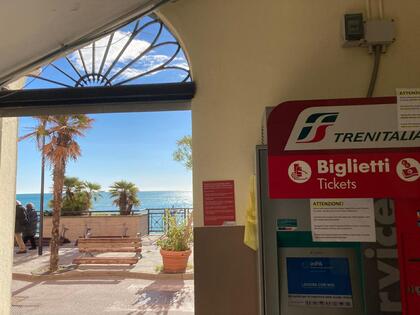 Biljettautomat från Trenitalia i en station med utsikt mot havet, palmer och bänkar utanför.