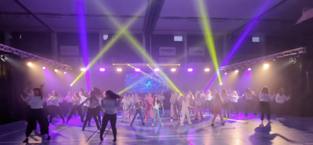 En stor grupp människor dansar på en scen med färgglada ljusstrålar i en inomhushall.