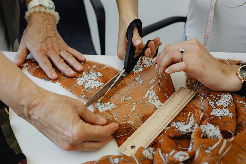 Två personer klipper ett blommigt tyg med sax och mäter med linjal på ett bord.