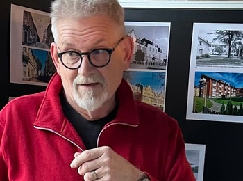 Två personer samtalar framför en utställningstavla med arkitekturbilder i en inomhusmiljö.