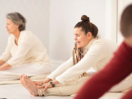 Två kvinnor sitter på golvet och gör stretchövningar i ett ljust rum, klädda i ljusa kläder.