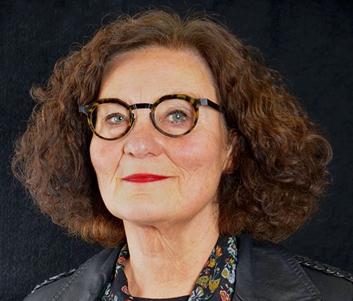 Person med lockigt brunt hår, svart läderjacka och blommig skjorta mot svart bakgrund.