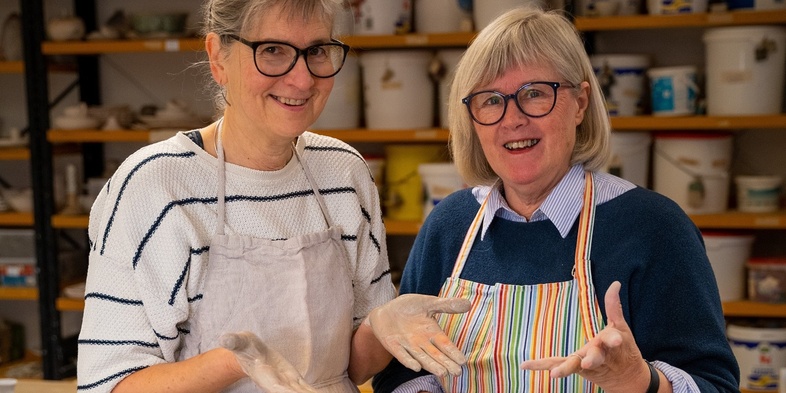 Två leende kvinnor med glasögon och förkläden står i en keramikverkstad med händerna täckta av lera.