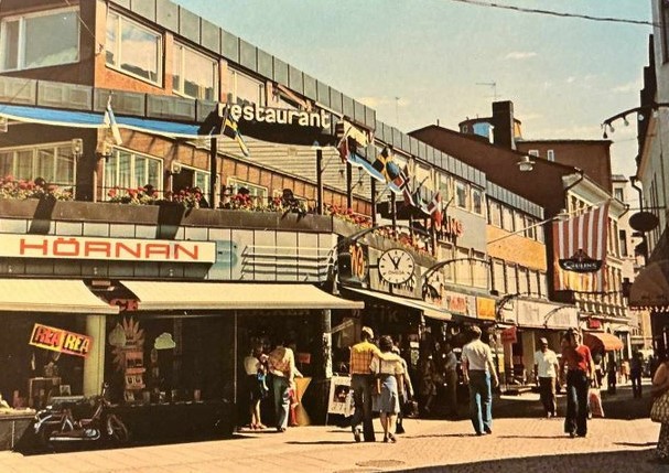 Gatuliv i stadsmiljö med butiker, restaurangskyltar, flaggor och människor som promenerar en solig dag.