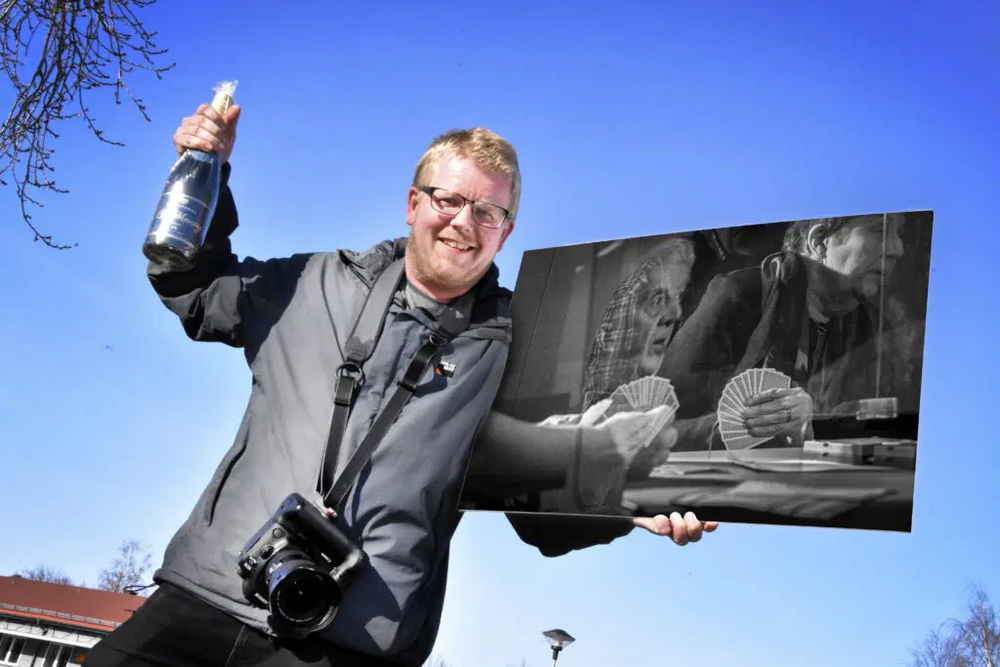 Person utomhus håller en champagneflaska och ett svartvitt foto, kamera hänger runt halsen.