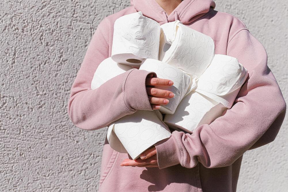 Person i rosa hoodie håller flera rullar toalettpapper mot en ljus vägg.