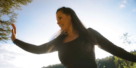 Person i svart tröja står utomhus med utsträckta armar mot en ljus himmel och skog i bakgrunden.