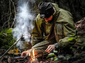 En person i friluftskläder tänder en eld i skogen med en vattenkokare hängande över elden.