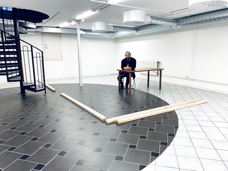 En person sitter vid ett bord i ett ljust rum med svart och vitt rutigt golv och spiraltrappa.