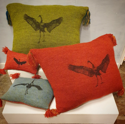 Fyra färgglada kuddar med fågelmotiv och tofsar, placerade på en vit piedestal.