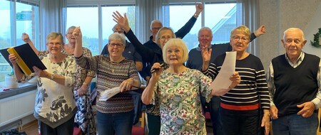 En grupp äldre personer sjunger tillsammans inomhus, flera håller papper och en kvinna har en mikrofon.