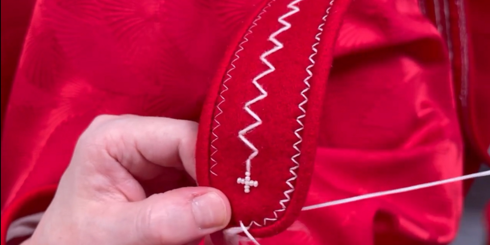 En hand broderar vita mönster på ett rött tyg med dekorativa sömmar och pärlor.