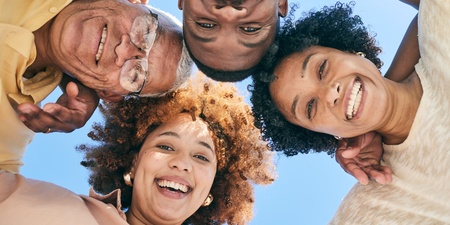 Fyra personer står i en cirkel och tittar ner mot kameran, alla ler mot varandra under en klar blå himmel.