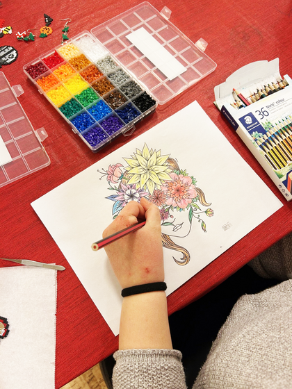 En person färglägger en teckning av ett ansikte med blommor i håret vid ett bord med färgpennor och pärlor.