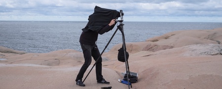 En person fotograferar med en stor kamera på stativ vid havet, täckt av en svart duk.