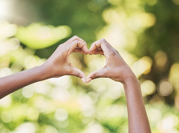 Två händer formar ett hjärta mot en suddig, grön bakgrund utomhus.