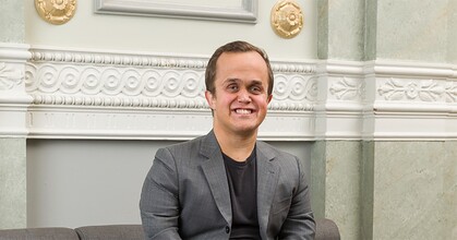 Man i grå kostym sitter på en soffa och ler, med dekorerad vägg i bakgrunden.