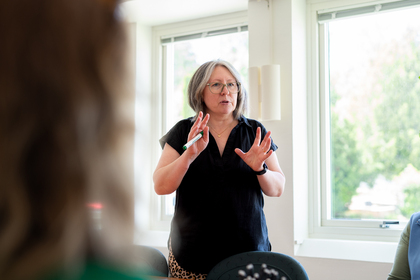 Kvinna med glasögon står och pratar framför ett fönster i ett ljust rum.