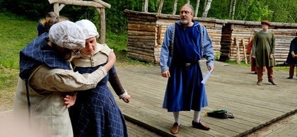 Personer i medeltida kläder spelar teater utomhus på en träscen med skog i bakgrunden.
