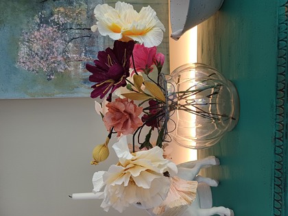 En glasvas med färgglada konstgjorda blommor står på ett turkost bord, med en tavla och dekorationer i bakgrunden.