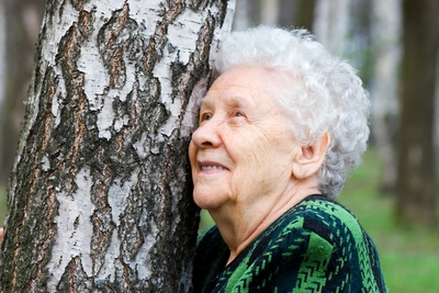 Äldre kvinna med vitt hår lutar sig mot ett träd i en skog och ler.