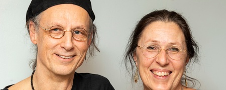 Ett leende par med glasögon står bredvid varandra mot en ljus bakgrund.